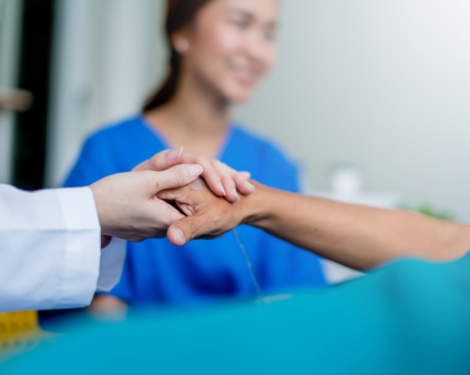 Primo piano di mani che stringono con cura e supporto quelle di un paziente, con un'operatrice sanitaria sorridente sfocata sullo sfondo - Fondo Sanità TLC